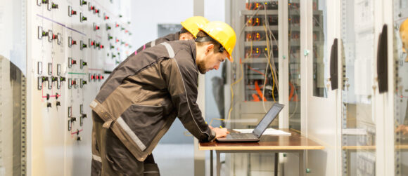 Engineers in hard hats standing next to an electrical control panel and looking at the screen of a laptop
