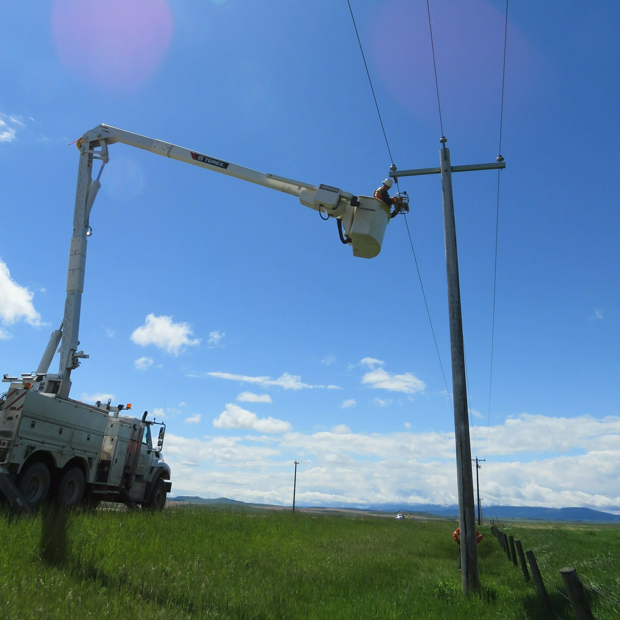 Outdoor photograph of a bucket truck lifting a worker within reach of a transmission tower
