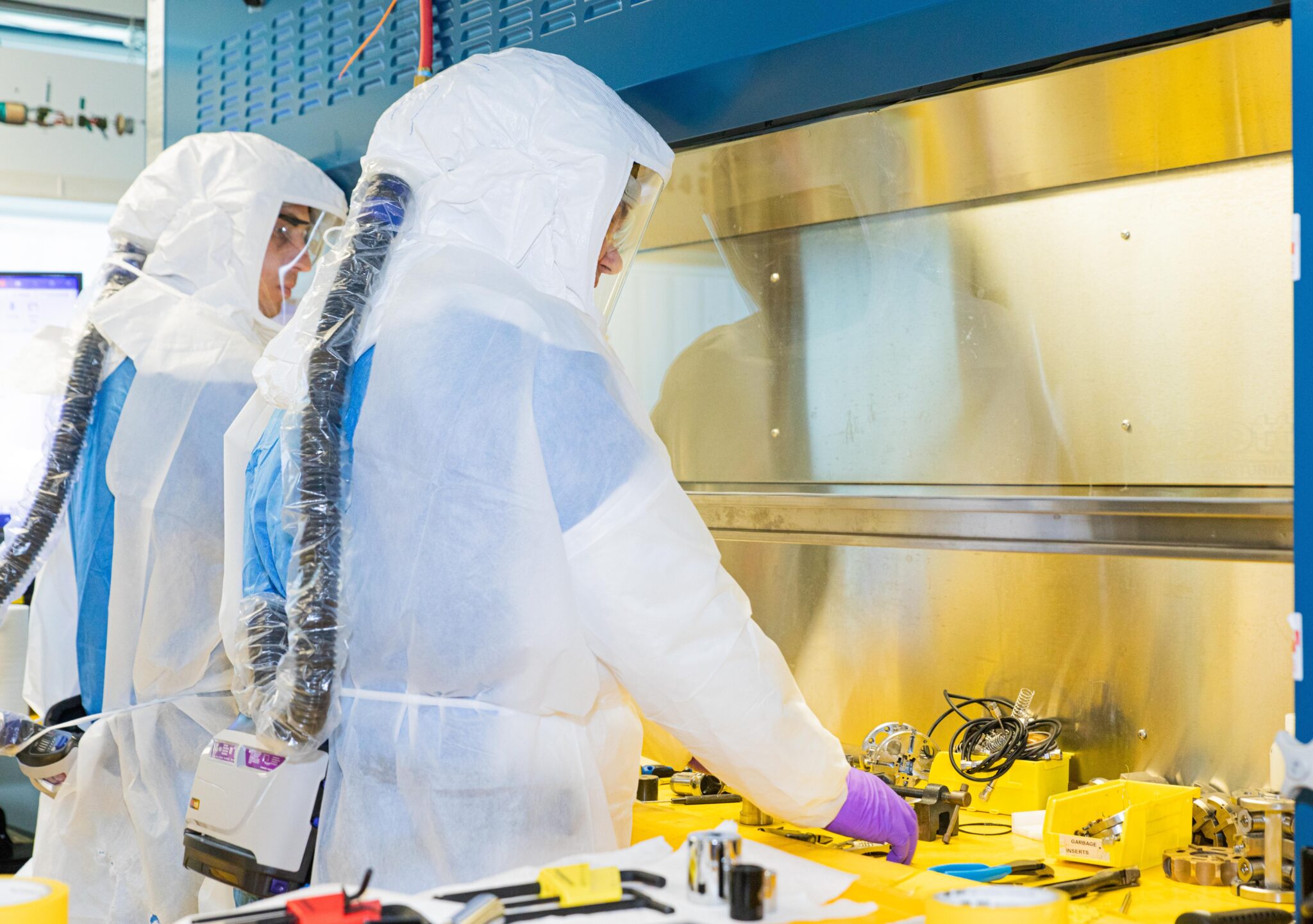 Two technicians in pressure suits servicing the Kinectrics Circumferential Wet Scrape Tool in a fume hood.
